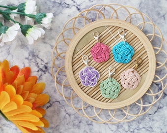 May include: Five colorful knotted flower charms with silver clasps on a bamboo tray. The charms are pink, blue, purple, green, and peach.