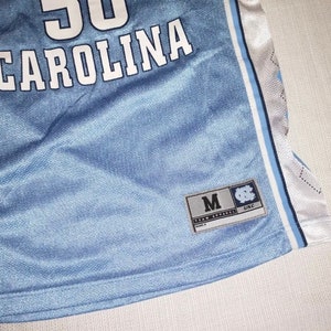 May include: A light blue basketball jersey with white lettering that says "CAROLINA" and a number "50". The jersey has white and light blue stripes on the sides. A tag on the bottom right corner says "M" for size and "UNC" for the University of North Carolina.