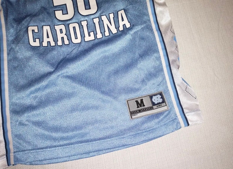 May include: A light blue basketball jersey with white lettering that says "CAROLINA" and a number "50". The jersey has white and light blue stripes on the sides. A tag on the bottom right corner says "M" for size and "UNC" for the University of North Carolina.