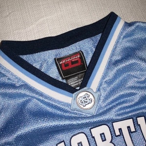 May include: A light blue basketball jersey with a white and light blue trim around the neck. The jersey has a white "NORTH" text on the front and a small, circular patch with the letters "UNC" in the center.