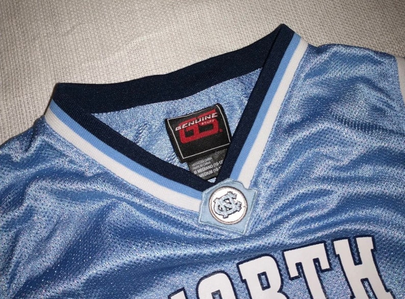 May include: A light blue basketball jersey with a white and light blue trim around the neck. The jersey has a white "NORTH" text on the front and a small, circular patch with the letters "UNC" in the center.