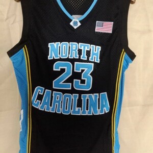 May include: Black basketball jersey with blue and yellow trim. The jersey has "NORTH" and "CAROLINA" in blue lettering and the number "23" in large blue lettering. A small American flag is on the right side of the jersey.