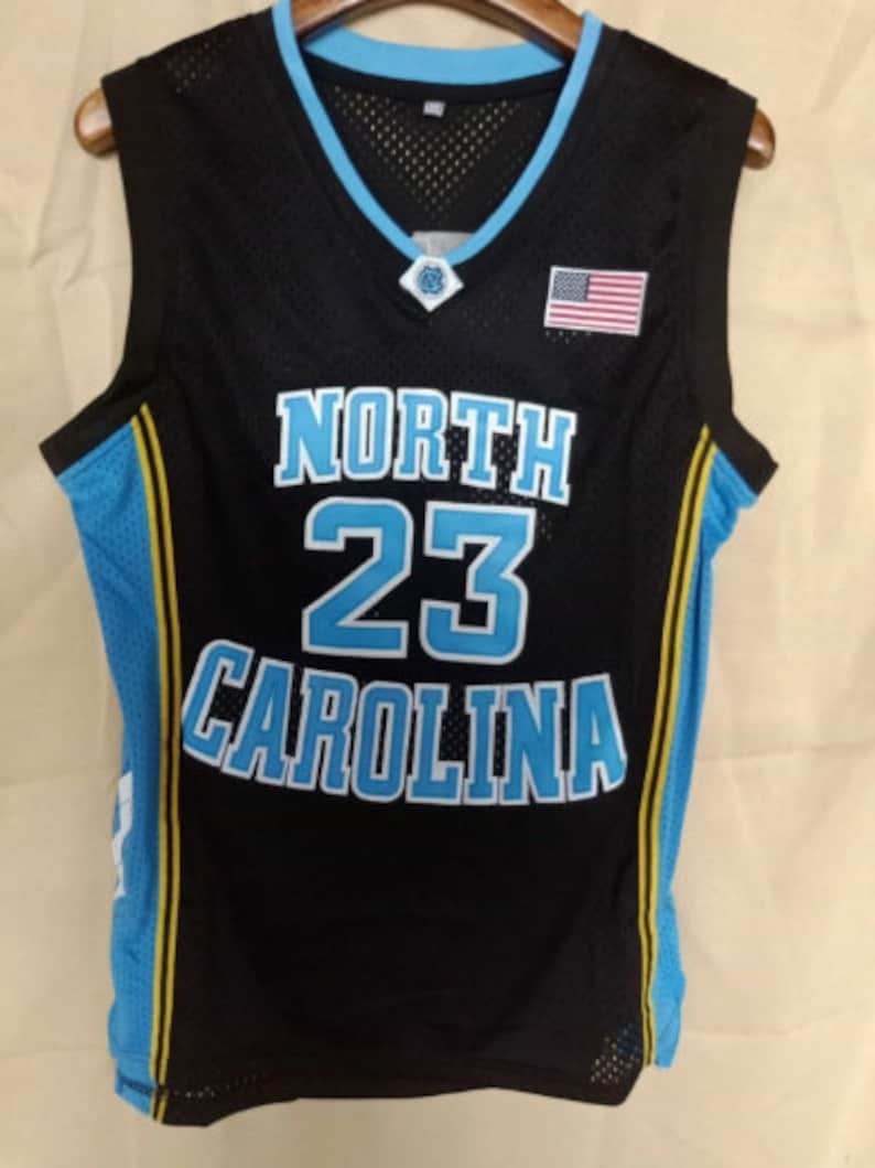May include: Black basketball jersey with blue and yellow trim. The jersey has "NORTH" and "CAROLINA" in blue lettering and the number "23" in large blue lettering. A small American flag is on the right side of the jersey.