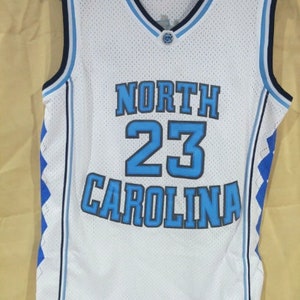 May include: White basketball jersey with blue and black trim. The jersey has the text "NORTH CAROLINA" and the number "23" on the front.