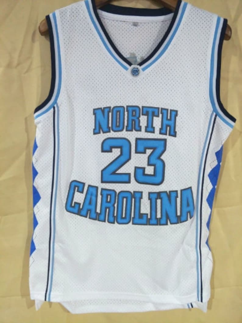 May include: White basketball jersey with blue and black trim. The jersey has the text "NORTH CAROLINA" and the number "23" on the front.