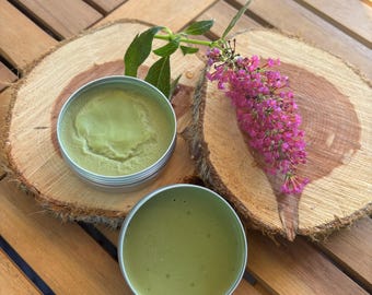 Könnte beinhalten: Zwei offene, silberfarbene Dosen mit grünem Balsam liegen auf einer Holzoberfläche. Eine Dose enthält einen strukturierten Balsam, die andere einen glatten Balsam. Ein Zweig mit Blättern und ein Büschel rosa Blüten sind ebenfalls vorhanden.