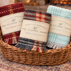 May include: Three miniature folded throw blankets in a woven wicker basket. Each blanket features a paper band labeled Little Cat Workshop The Classic Throw, with color variants Mulberry Plaid, Heritage Navy, and Seafoam Plaid with fringe.