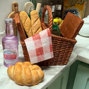 May include: A miniature picnic scene featuring a woven basket with bread, a red and white checkered napkin, and a bottle of rose lemonade. A small loaf of bread sits in front of the basket. Other items include a small green bottle and a brown book.