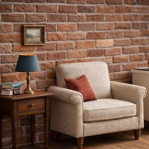 May include: Miniature living room scene with a beige armchair, a small wooden side table with a lamp, and a miniature stove. A red brick wall serves as the backdrop, with a small framed picture above the chair. The lamp has a blue shade.