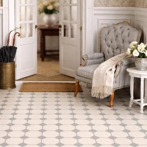 May include: A cozy interior scene featuring a wooden side table with a lamp, books, and a potted plant. A tufted armchair with a blanket sits near a small table with flowers. The room has patterned flooring and white doors.