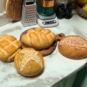 May include: A selection of miniature bread loaves and rolls. The bread is a golden brown colour, with different textures, including a lattice pattern and a rustic crust. One loaf sits on a small wooden chopping board. The scene suggests a bakery or food-themed display.