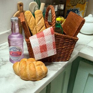 May include: A miniature picnic scene featuring a woven basket with bread, a red and white checked napkin, and a bottle of rose lemonade. A croissant and other food items are on the counter. The scene is set on a white and grey countertop.