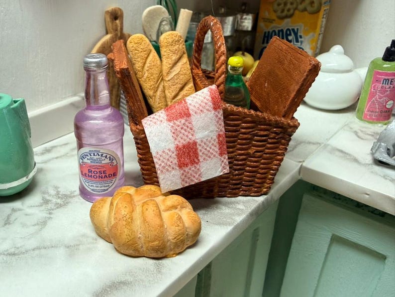May include: A miniature picnic scene featuring a woven basket with bread, a red and white checked napkin, and a bottle of rose lemonade. A croissant and other food items are on the counter. The scene is set on a white and grey countertop.