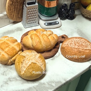 May include: A selection of miniature baked goods, including a cross-scored loaf, a braided loaf on a wooden board, a country loaf, and a rustic roll. The breads are golden brown and rest on a white surface.