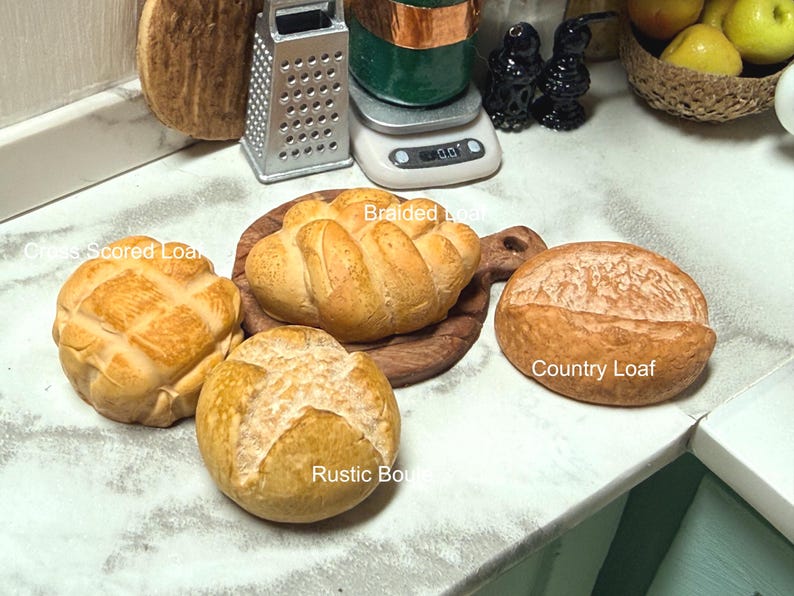 May include: A selection of miniature baked goods, including a cross-scored loaf, a braided loaf on a wooden board, a country loaf, and a rustic roll. The breads are golden brown and rest on a white surface.