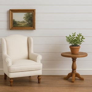 May include: A miniature interior scene featuring a cream-colored armchair, a small round wooden side table with a potted plant, and a framed landscape painting. The scene is set against a white paneled wall and a wooden floor.