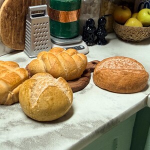 May include: A selection of miniature bread rolls, including a round loaf and several crusty rolls with textured surfaces. The bread is a golden brown colour, suggesting a fresh bake. The scene is set in a kitchen environment, with a grater and other kitchen items in the background.