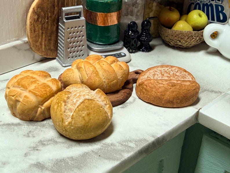 May include: A selection of miniature bread rolls, including a round loaf and several crusty rolls with textured surfaces. The bread is a golden brown colour, suggesting a fresh bake. The scene is set in a kitchen environment, with a grater and other kitchen items in the background.