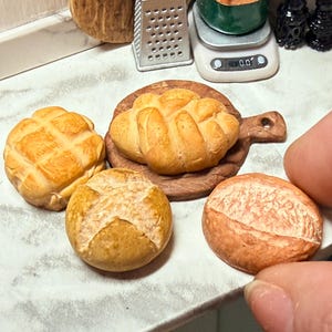 May include: A collection of miniature baked goods, including a round loaf, a bread roll, and a loaf on a wooden board. The bread has a golden-brown crust and detailed textures. The scene is set on a white countertop.