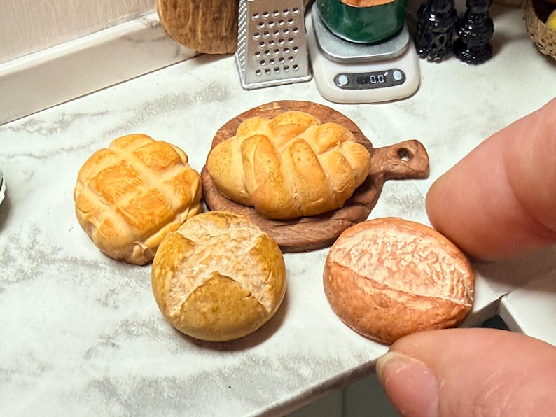 May include: A collection of miniature baked goods, including a round loaf, a bread roll, and a loaf on a wooden board. The bread has a golden-brown crust and detailed textures. The scene is set on a white countertop.