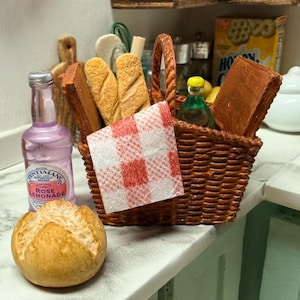 May include: A miniature picnic scene with a woven basket containing bread, a red and white checked napkin, and a bottle of rose lemonade. A loaf of bread and a green bottle are also present, creating a charming, small-scale picnic setting.