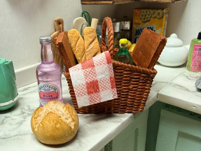 May include: A miniature picnic scene with a woven basket containing bread, a red and white checked napkin, and a bottle of rose lemonade. A loaf of bread and a green bottle are also present, creating a charming, small-scale picnic setting.