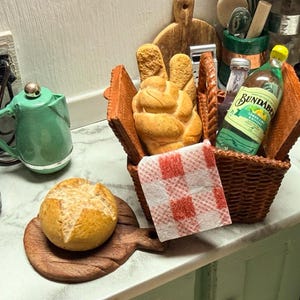 May include: A miniature picnic scene featuring a woven basket with bread, a bottle of Bundaberg Lemon & Bitters, and a red and white checkered napkin. A small loaf of bread rests on a wooden board. A green kettle is also visible.
