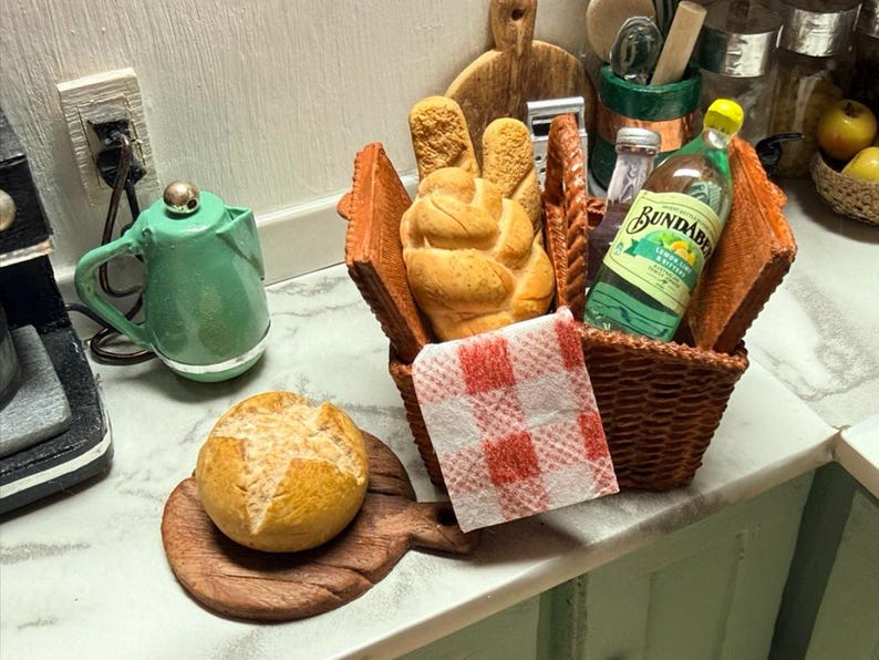 May include: A miniature picnic scene featuring a woven basket with bread, a bottle of Bundaberg Lemon & Bitters, and a red and white checkered napkin. A small loaf of bread rests on a wooden board. A green kettle is also visible.