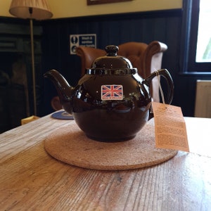 Può includere: Una teiera in ceramica marrone scuro con coperchio e manico, appoggiata su un sottobicchiere di sughero. La teiera ha un adesivo con la bandiera britannica e la scritta "MADE IN ENGLAND". Un'etichetta è appesa.