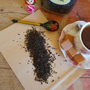 May include: A brown paper bag with loose black tea leaves, a wooden spoon with a painted design, a small porcelain doll, a jar of tea labeled "Russian Caravan", and a porcelain teacup with a saucer and caramel candies.