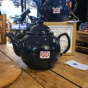 May include: Two navy blue ceramic teapots with lids and handles. Each teapot has a Union Jack sticker that reads "Made in England." One teapot is in front of a box that reads "Adderley Ceramics." The teapots are on a wooden table.