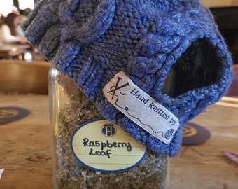 Op de afbeelding: Een blauwe gebreide muts met een label met de tekst "Hand knitted by" en een pot met losse bladthee met het label "Raspberry Leaf".