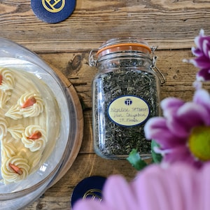 May include: A glass jar filled with loose leaf green tea. The jar has a blue label with the text "Nepalese SFTGFOP1  First Flush" and a logo of a letter H inside a circle. The jar is sitting on a wooden table with a pink flower in the foreground.