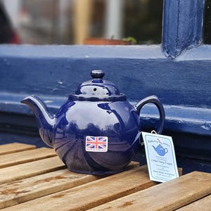 Op de afbeelding: Marineblauwe keramische theepot met een Union Jack-sticker en een label van Adderley Ceramics. De theepot staat op een houten tafel. Het label luidt "Cobalt Berry Teapot".
