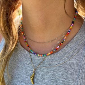 Könnte beinhalten: Ein mehrschichtiges Halsketten-Set mit einer Perlenkette mit bunten Perlen, einer filigranen Silberkette und einem goldenen Anhänger. Die Halsketten werden von einer Person getragen, die ein graues T-Shirt trägt.