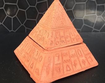 Könnte beinhalten: Eine terracottafarbene, pyramidenförmige Box mit Deckel. Die Box ist mit geschnitzten Hieroglyphen und geometrischen Mustern verziert. Die Pyramide ist ein dekorativer Gegenstand, wahrscheinlich zur Aufbewahrung kleiner Gegenstände.