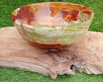 May include: A handcrafted, polished stone bowl with a natural, organic shape. The bowl features a blend of red, green, and brown hues, creating a unique, marbled appearance. It rests on a piece of weathered wood against a backdrop of green grass.