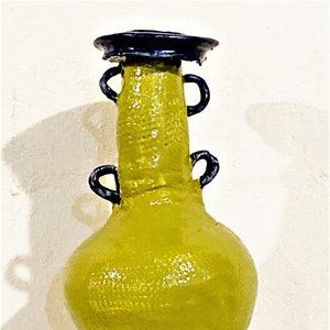 May include: A green ceramic vase with a brown base and blue handles. The vase has a textured surface and a wide, round body.