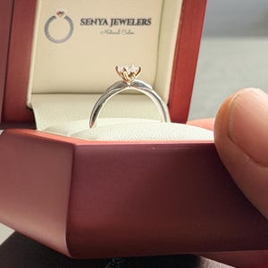 May include: A diamond engagement ring in a red jewelry box. The ring features a silver band and a gold setting holding a clear diamond. The box is open, revealing a white interior and the Senya Jewelers logo.