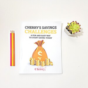 May include: A white booklet with the title "Cherry's Savings Challenges: A fun and easy way to start saving today."  The cover features a cartoon illustration of a gold money bag with a dollar sign and a stack of gold coins. The text "Life of Cherry" is at the bottom of the cover.