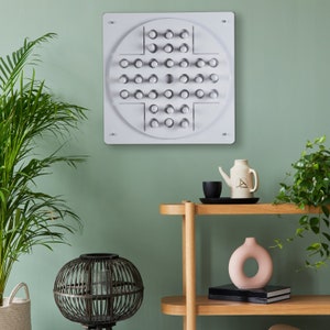 May include: White wooden wall mounted game board with a circular design and 49 holes. The board is mounted on a light green wall. A wooden shelf with a vase, a teapot, and a cup is below the board.