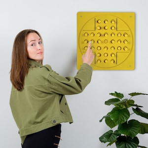 Peut inclure: Une personne interagit avec un jeu mural jaune avec une grille circulaire de boules jaunes. Le jeu ressemble à un jeu de pions classique.