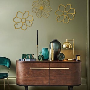 May include: Three gold metal wall decor pieces in the shape of flowers. The flowers are on a sage green wall above a dark wood dresser with gold hardware. The dresser has a green velvet chair in front of it.