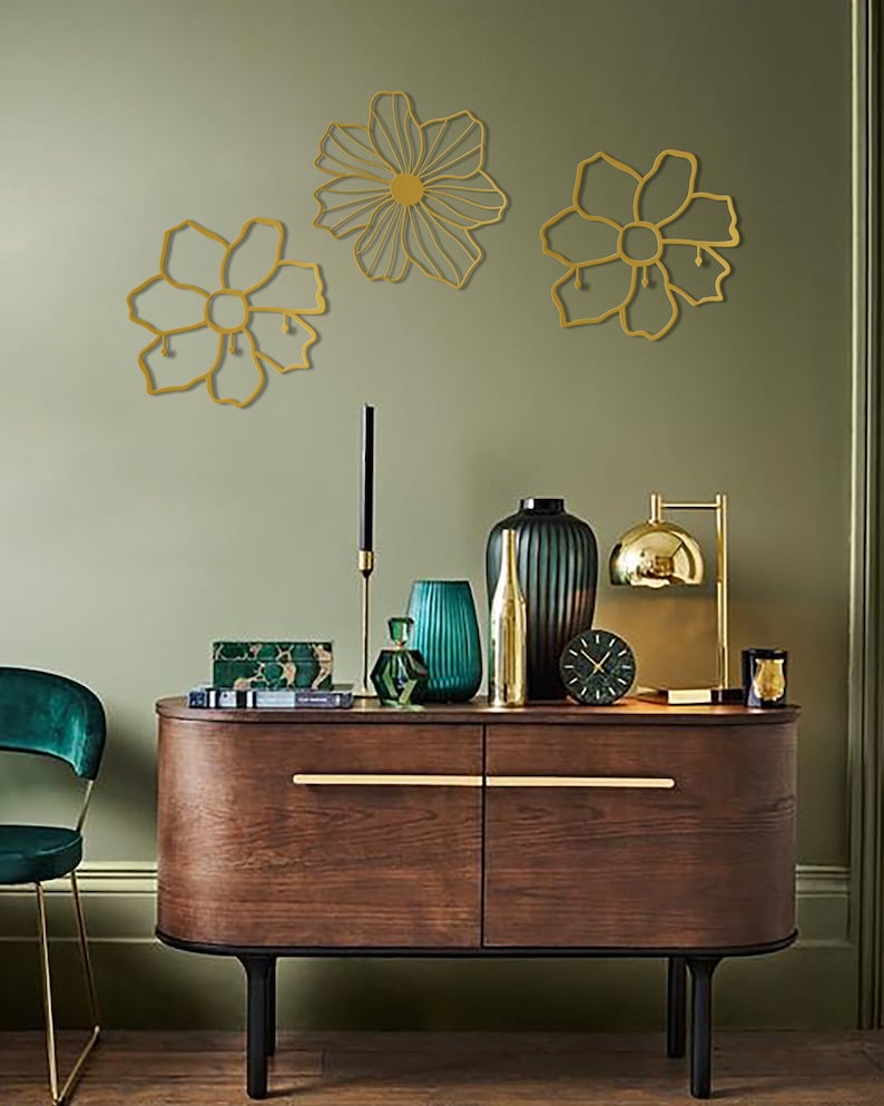 May include: Three gold metal wall decor pieces in the shape of flowers. The flowers are on a sage green wall above a dark wood dresser with gold hardware. The dresser has a green velvet chair in front of it.