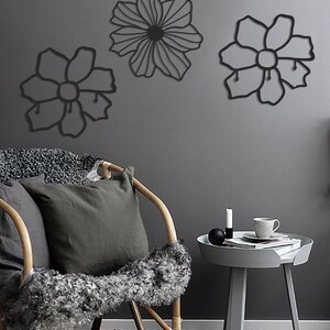 May include: Three black metal wall decor pieces in the shape of flowers. The flowers are attached to a gray wall. A wicker chair with a gray pillow and a gray side table with a cup and saucer are in the foreground.