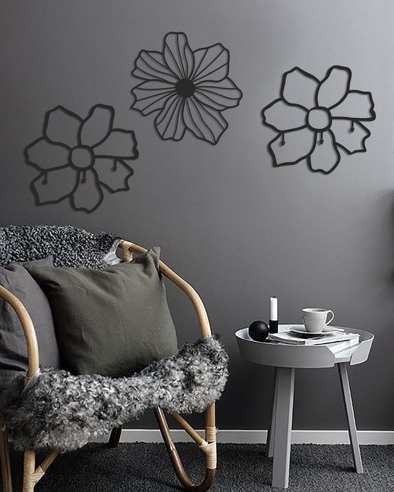May include: Three black metal wall decor pieces in the shape of flowers. The flowers are attached to a gray wall. A wicker chair with a gray pillow and a gray side table with a cup and saucer are in the foreground.