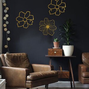 May include: Three gold metal wall decor pieces in the shape of flowers. The flowers are outlined in gold and have a simple design. The flowers are mounted on a dark gray wall.