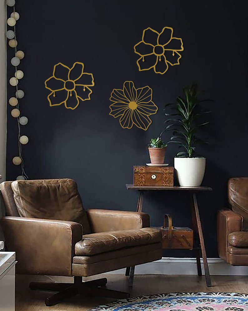 May include: Three gold metal wall decor pieces in the shape of flowers. The flowers are outlined in gold and have a simple design. The flowers are mounted on a dark gray wall.