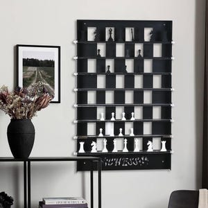 May include: Black wall-mounted chessboard with white and black squares. The chessboard has shelves for storing chess pieces. The chess pieces are white and black plastic. The chessboard has the word "CHESS" written on the bottom.