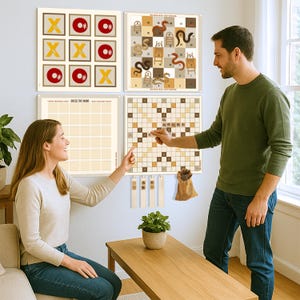 May include: Four framed game boards are displayed on a wall, including tic-tac-toe, a snakes and ladders game, a crossword puzzle, and a "Guess the Word" board. A couple points at the crossword puzzle. A small plant sits on a wooden table.